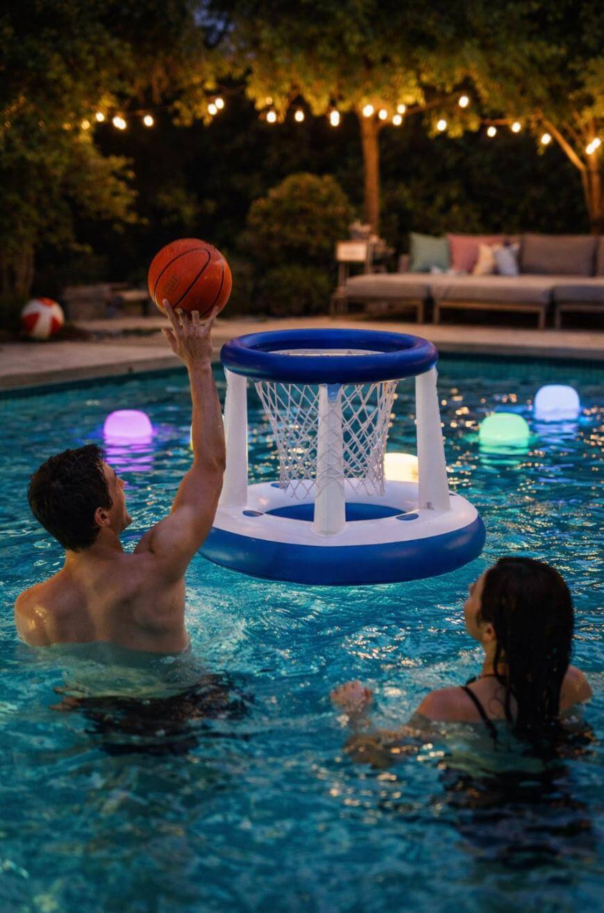 A man in a pool lifts a basketball toward a floating inflatable basketball hoop, while a woman watches. Colorful lights float nearby, and string lights hang above a patio with outdoor seating in the background.