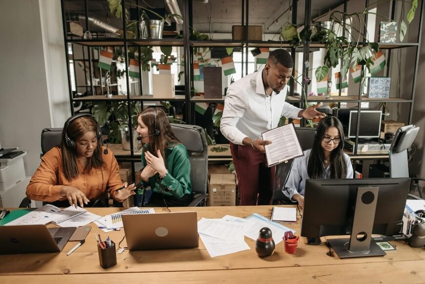 Four coworkers sit and work at a shared desk in a modern office. One man is standing and gesturing with papers, while three women are seated at computers, discussing and working on documents. The background is decorated with plants.