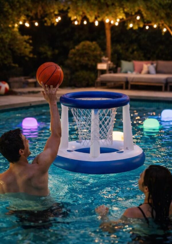 A man in a pool prepares to shoot a basketball into an inflatable hoop while a woman watches. Colorful floating lights and string lights decorate the pool area, with patio furniture in the background.