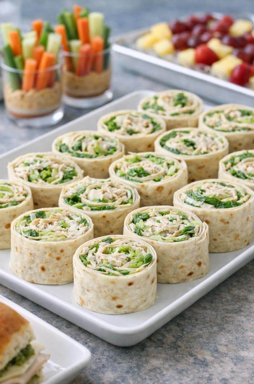 A platter of tortilla pinwheel sandwiches filled with greens and shredded chicken is in focus. In the background are cups of veggie sticks with dip and a tray of fruit skewers. The food is arranged on a gray surface.