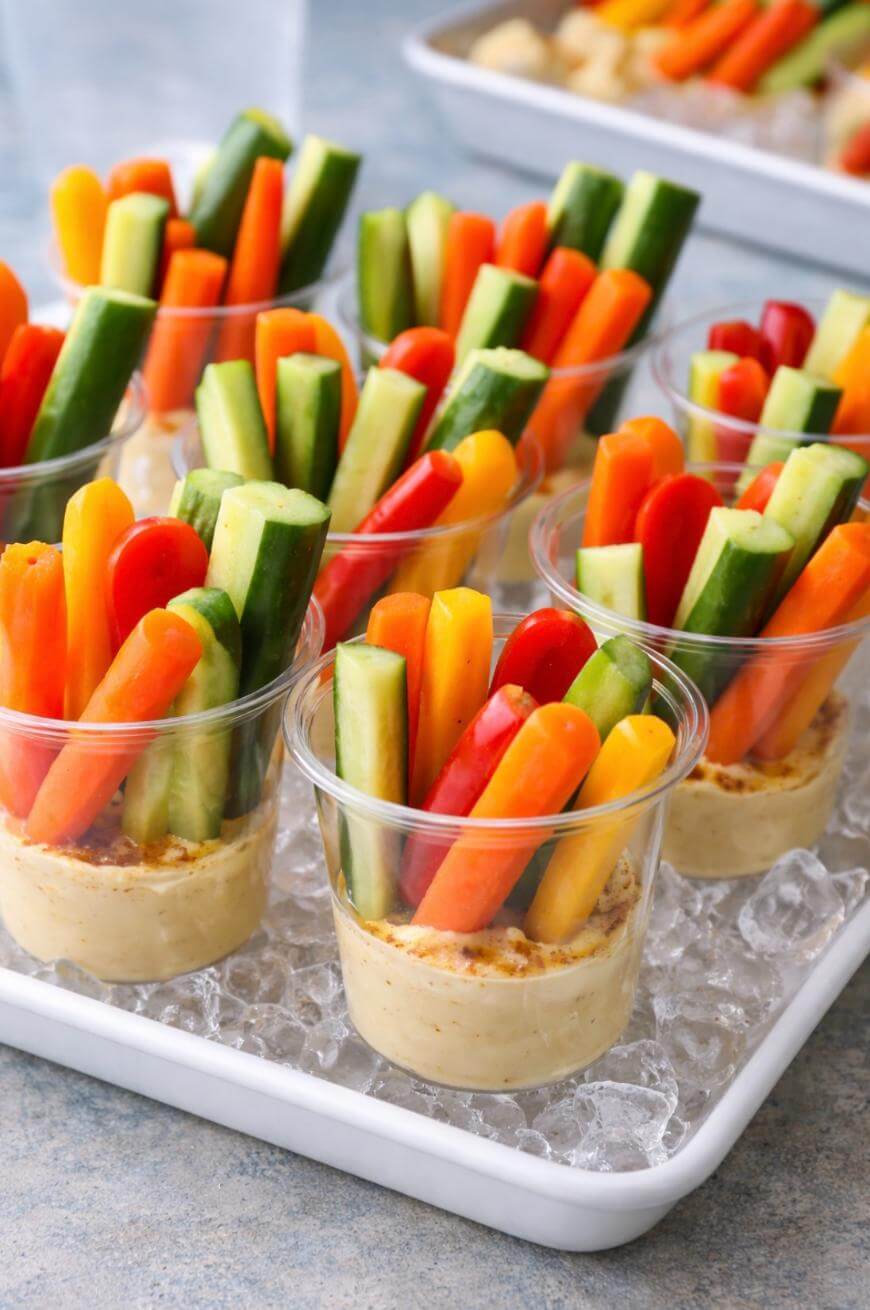 Clear plastic cups filled with hummus and assorted fresh vegetable sticks, including cucumbers, carrots, and red bell peppers, are arranged on a tray of ice.
