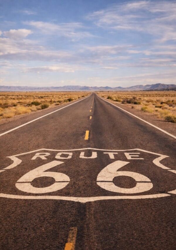 A straight, empty highway stretches into the distance through a desert landscape, with Route 66 painted in large white letters on the road under a blue sky with scattered clouds.