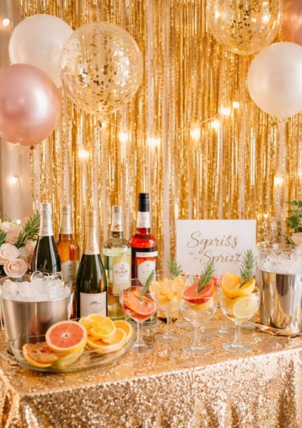 A sparkling party table with bottles of wine, cocktails garnished with fruit, ice buckets, sliced citrus, flowers, and gold, white, and pink balloons, set against a gold tinsel backdrop with string lights.