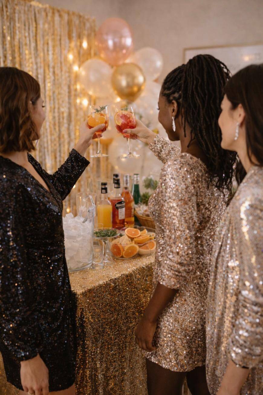 Three women dressed in sparkly dresses toast with colorful cocktails at a festive party table decorated with gold, balloons, bottles, and sliced oranges.