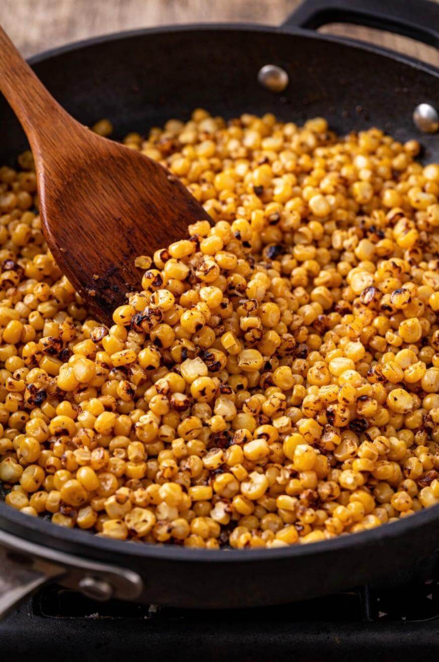 A wooden spoon stirs roasted corn kernels in a black skillet, showing golden-brown, slightly charred pieces.