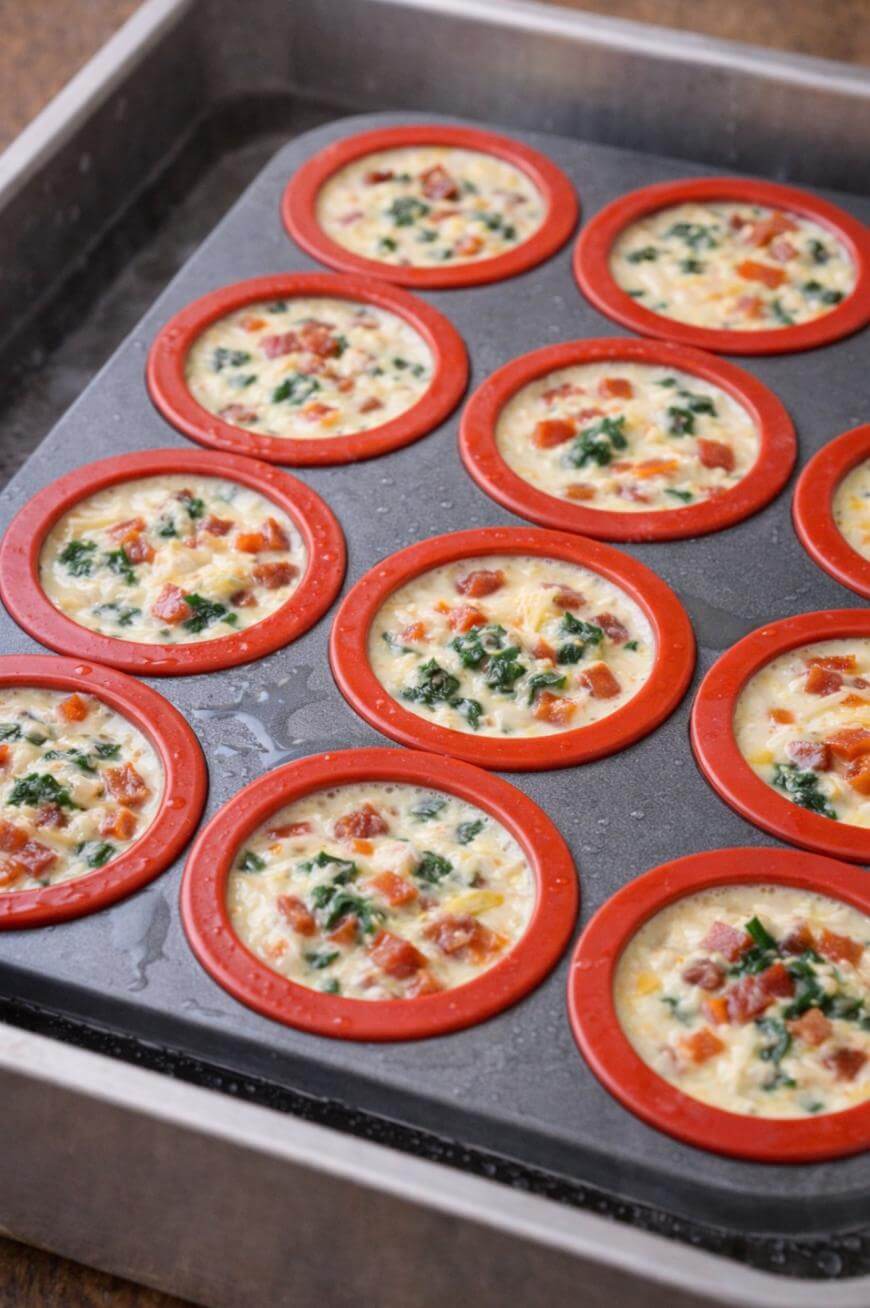 A baking tray with multiple circular silicone molds filled with a mixture of eggs, vegetables, and herbs, ready to be baked into mini frittatas or egg muffins.
