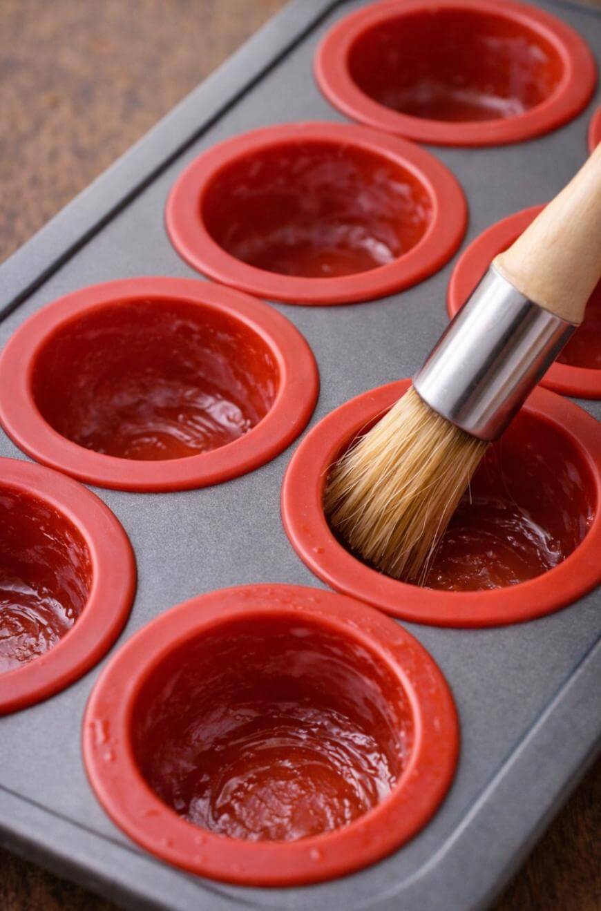 A muffin tin with red silicone liners is being brushed with oil or butter using a pastry brush, preparing it for baking.