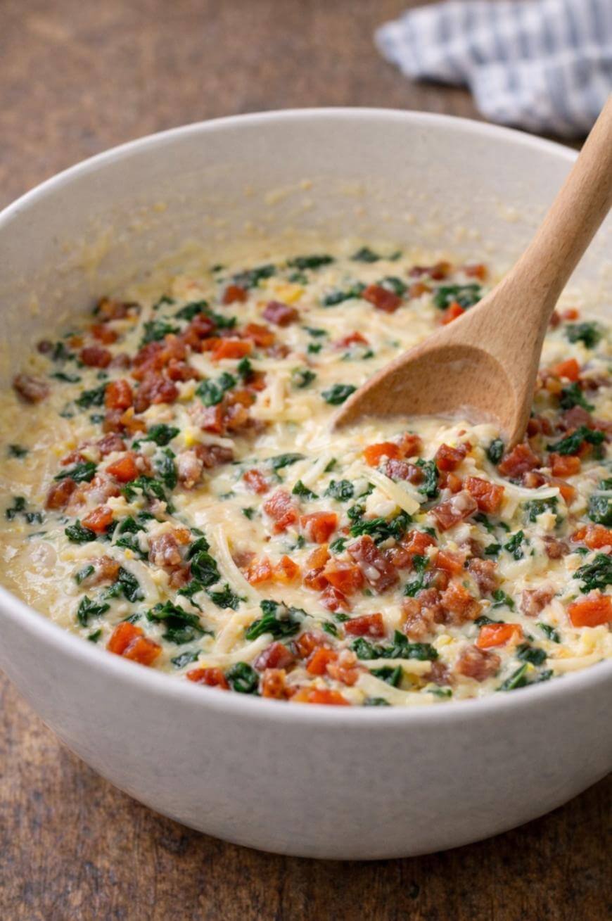 A large white mixing bowl filled with a creamy mixture of chopped spinach, shredded cheese, and diced bacon, being stirred with a wooden spoon.