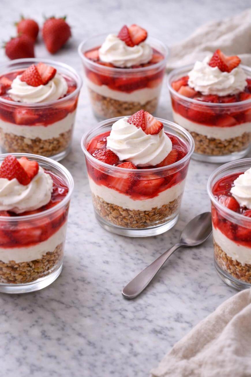Six glass cups filled with layered strawberry parfaits, topped with whipped cream and fresh strawberry slices, are arranged on a marble surface with a spoon and fresh strawberries nearby.