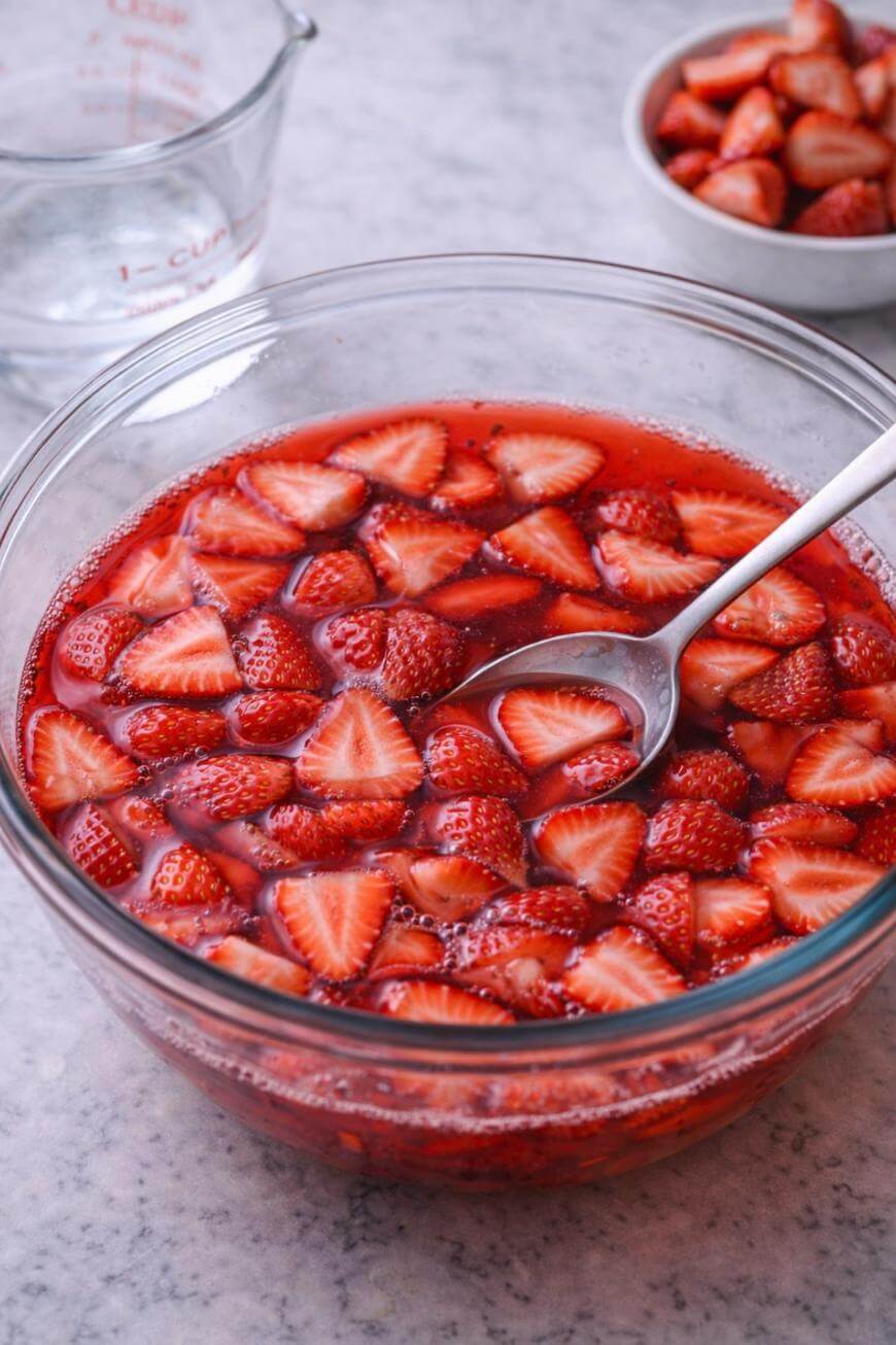 A glass bowl filled with sliced strawberries in syrup, with a metal spoon resting inside. In the background are a measuring cup with liquid and a small bowl of more strawberry slices.