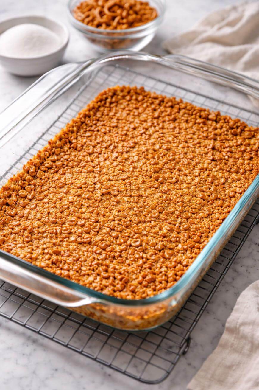 A glass baking dish filled with golden-brown cereal bars sits on a cooling rack. Two bowls in the background contain sugar and cereal, and a light cloth is partially visible on the side.