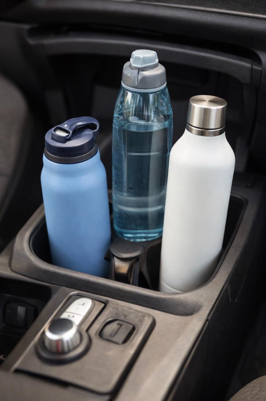 Three reusable water bottles—one blue, one transparent with a gray cap, and one white with a silver cap—are placed in the cup holders of a car’s center console.