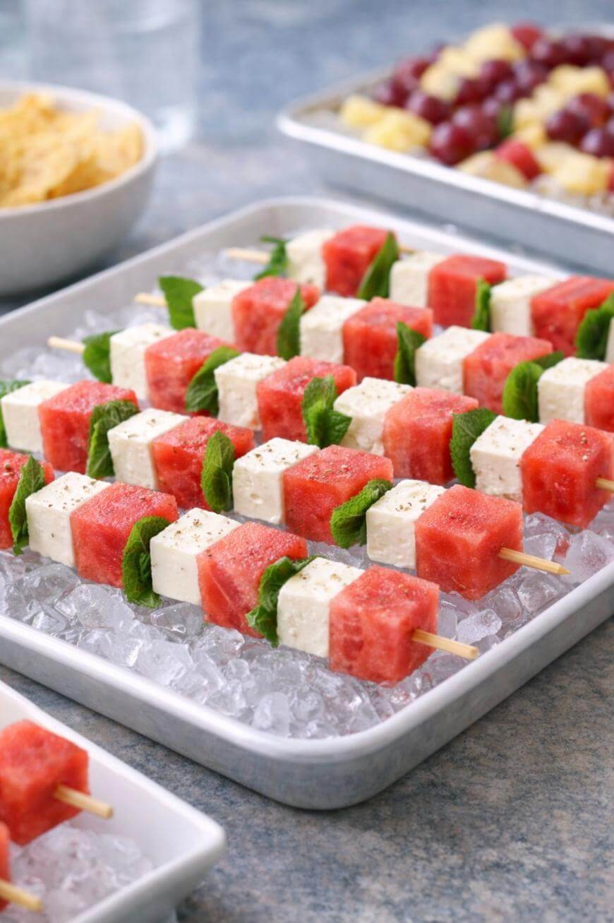 Wooden skewers with alternating cubes of watermelon, feta cheese, and fresh mint leaves are arranged neatly on a tray of ice. Other trays with different skewers are in the background.