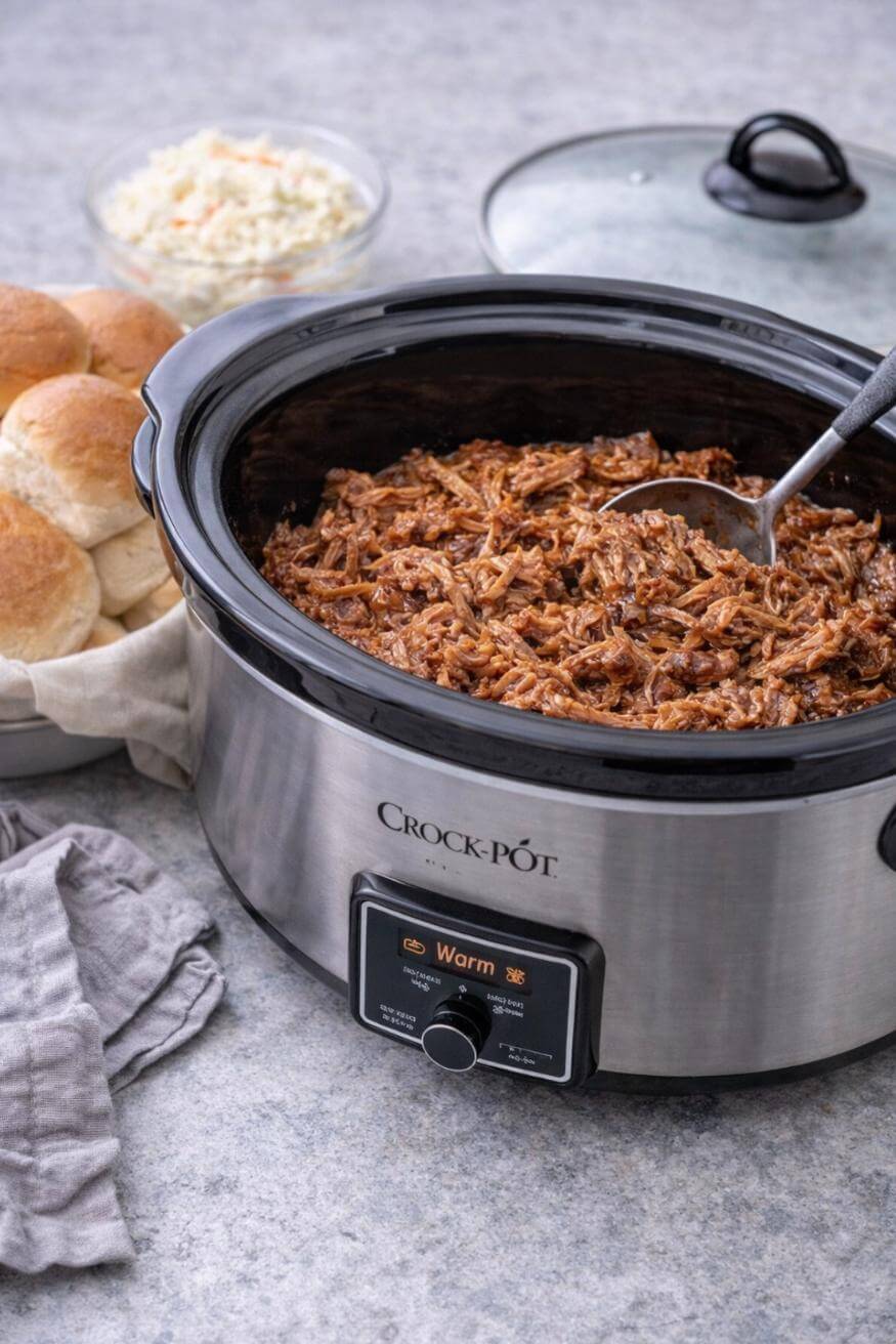 A Crock-Pot filled with shredded barbecue meat sits on a counter, with a serving spoon inside. Nearby are a plate of dinner rolls, a bowl of coleslaw, and a gray napkin. The Crock-Pot lid rests to the side.