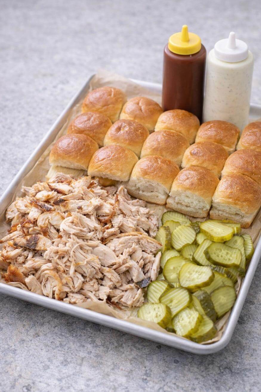 A tray with rows of slider rolls, shredded chicken, sliced pickles, and two squeeze bottles of barbecue and creamy white sauce on a light gray surface.