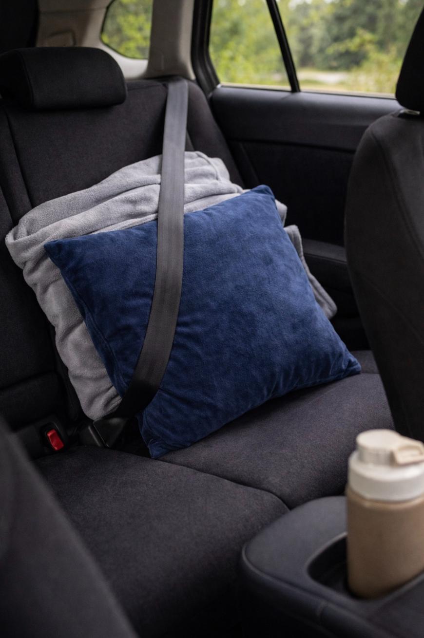 A blue pillow and a folded gray blanket are secured with a seatbelt on the backseat of a car. A reusable cup sits in a cupholder in the foreground. Greenery is visible outside the car window.