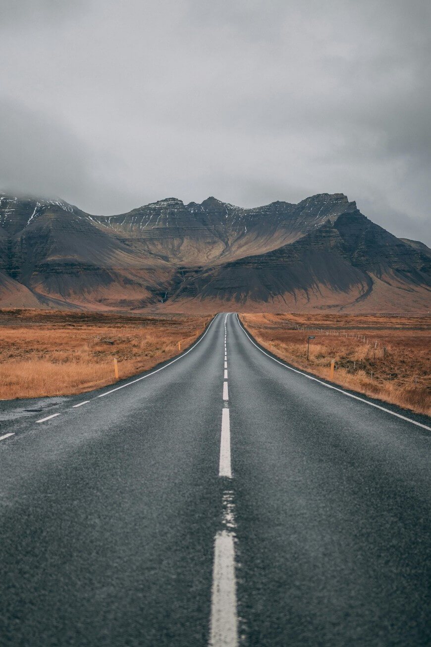 A straight, empty road stretches into the distance, leading toward rugged mountains under a cloudy sky, with brown grassland on either side of the road.