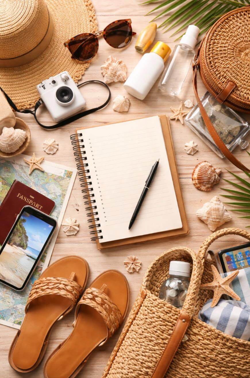 Flat lay of summer travel essentials including a straw hat, sunglasses, camera, sunscreen, notebook with pen, seashells, sandals, woven bag, passport, map, phone, and water bottle on a light wooden surface.