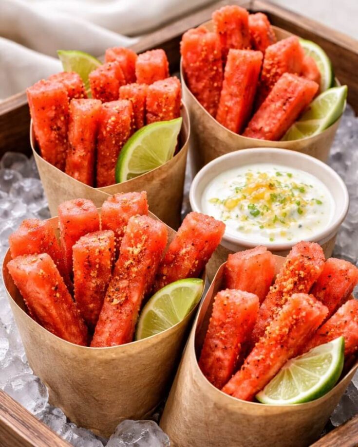 Four brown paper cones filled with seasoned, red watermelon sticks and lime wedges, arranged around a bowl of creamy dipping sauce, all set on a tray of ice.