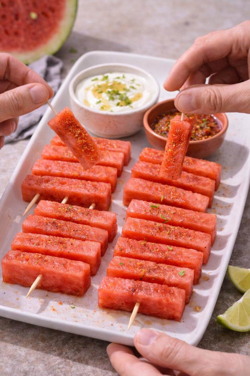 Rectangular watermelon sticks on skewers are arranged on a platter, garnished with seasoning. Two hands pick up pieces. Dips in small bowls and lime wedges are on the tray, with part of a watermelon in the background.
