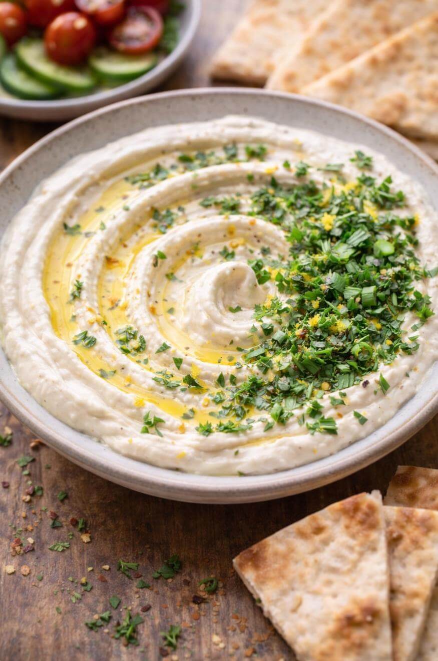 A bowl of creamy dip swirled with olive oil and topped with chopped green herbs, served with pita bread on the side. Sliced cucumber and cherry tomatoes are visible in the background.