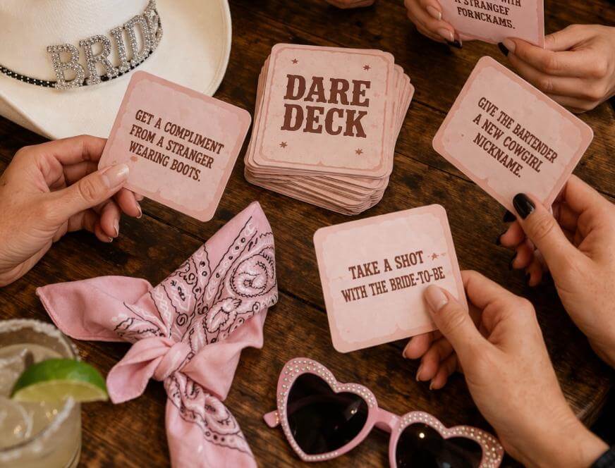 Four women sit around a wooden table with a “Dare Deck” card game. Visible cards read various dares. Also on the table are a cocktail, pink bandana, pink heart-shaped sunglasses, and a white hat labeled “Bride.”.
