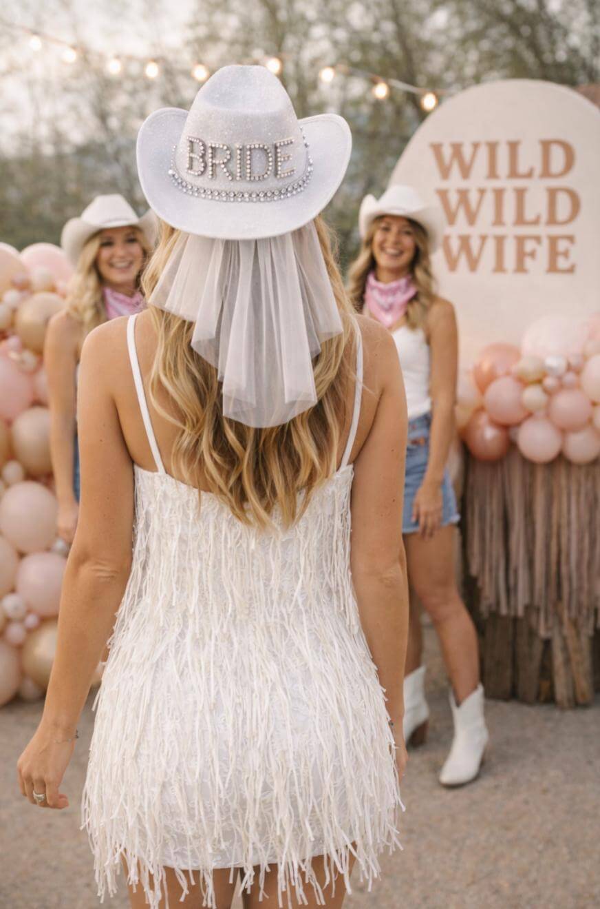 A woman in a white dress and BRIDE cowboy hat stands with her back to the camera at a bachelorette party, facing two women in western outfits. A “WILD WILD WIFE” sign and pink balloons are in the background.