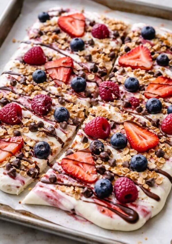 A tray of yogurt bark topped with strawberries, raspberries, blueberries, granola, mini chocolate chips, and a drizzle of chocolate, cut into rectangular pieces.