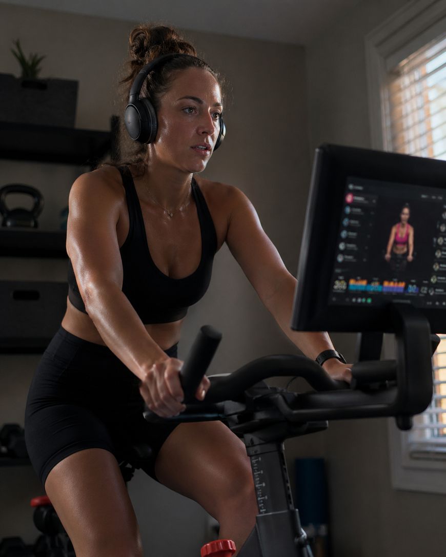 A woman wearing headphones and black workout clothes rides an exercise bike indoors, focused on a screen displaying a virtual cycling class. Shelves with gym equipment and a window are visible in the background.