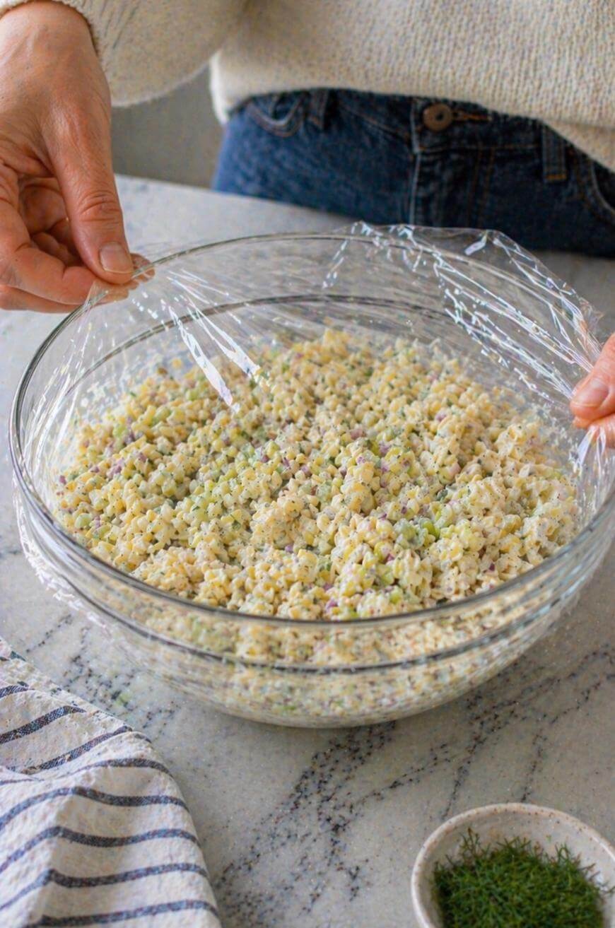 A person covers a glass bowl filled with mixed couscous and chopped vegetables using plastic wrap on a kitchen counter. A striped towel and a small bowl of herbs are nearby.