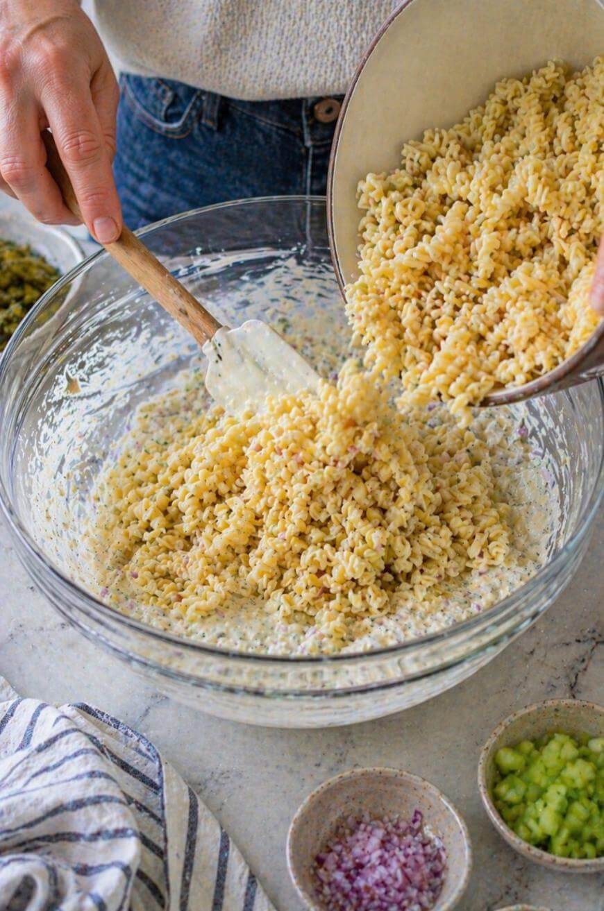 A person pours cooked pasta from a bowl into a larger glass bowl of creamy sauce, using a spatula. Small bowls of chopped red onion and celery are on the counter nearby.