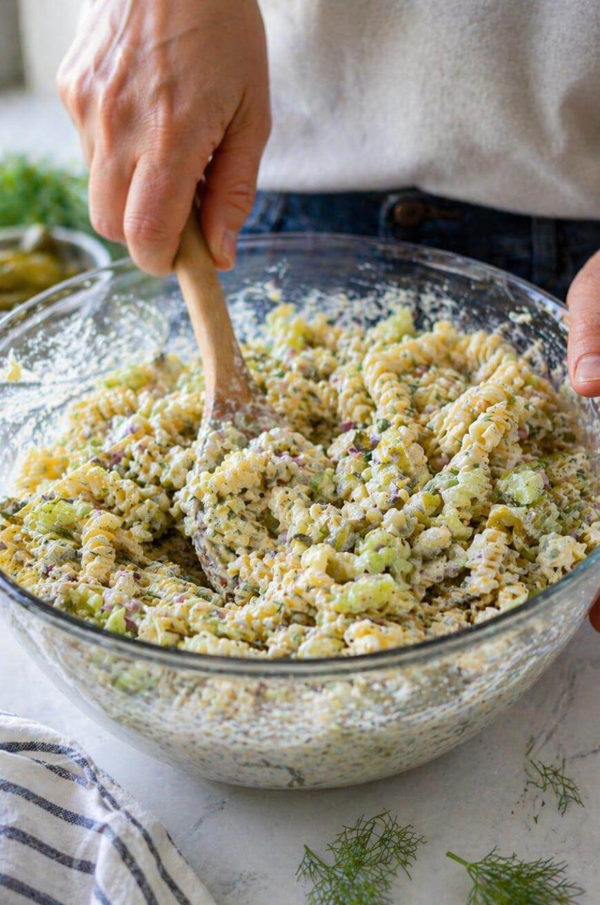 A person stirs a creamy pasta salad with rotini noodles, chopped celery, and herbs in a large glass bowl using a wooden spoon. A striped cloth and fresh dill are visible nearby on the countertop.