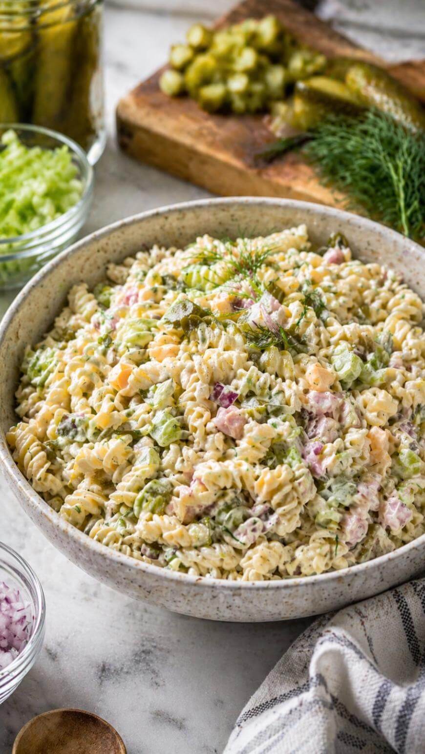 A bowl of creamy pasta salad with rotini noodles, diced vegetables, and herbs sits on a table. Chopped pickles and celery are visible in bowls and on a cutting board in the background.
