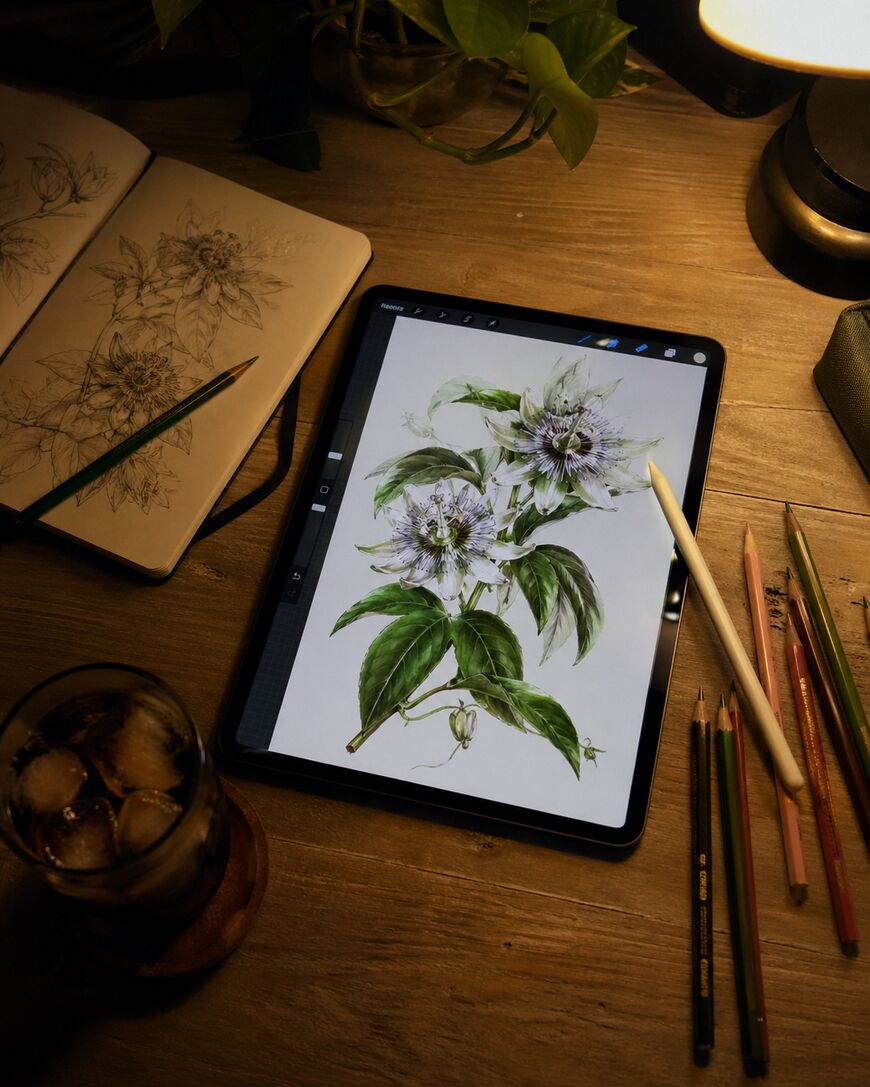 A digital tablet on a wooden desk displays a detailed botanical illustration. Nearby are colored pencils, a sketchbook with similar drawings, a drink with ice, and a lit lamp, creating a cozy creative workspace.