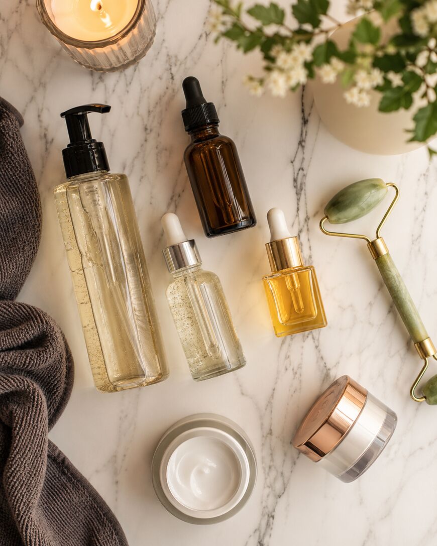 Flat lay of skincare items on a marble surface: a candle, brown towel, glass bottles with droppers, pump bottle, face cream jar, a jade roller, and a vase with white flowers. Spa and self-care theme.