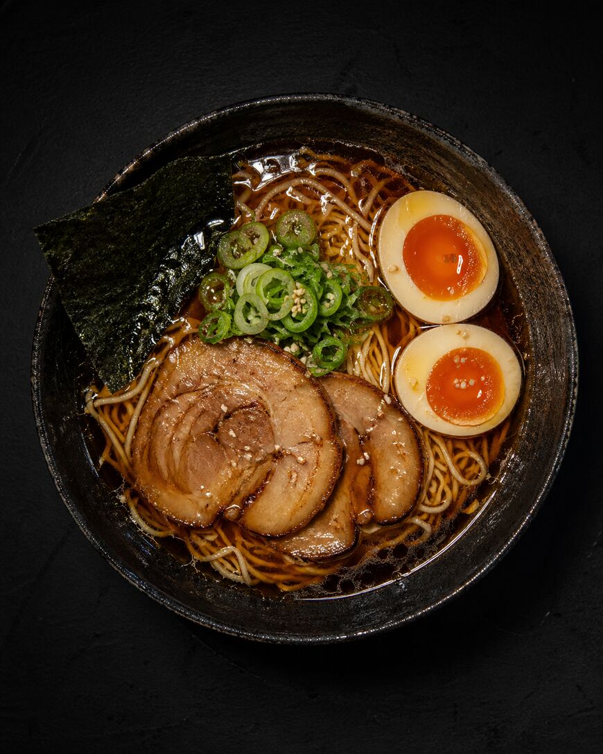 A bowl of ramen with sliced pork, halved soft-boiled eggs, green onions, seaweed, and noodles in a dark broth, served in a black bowl on a dark background.