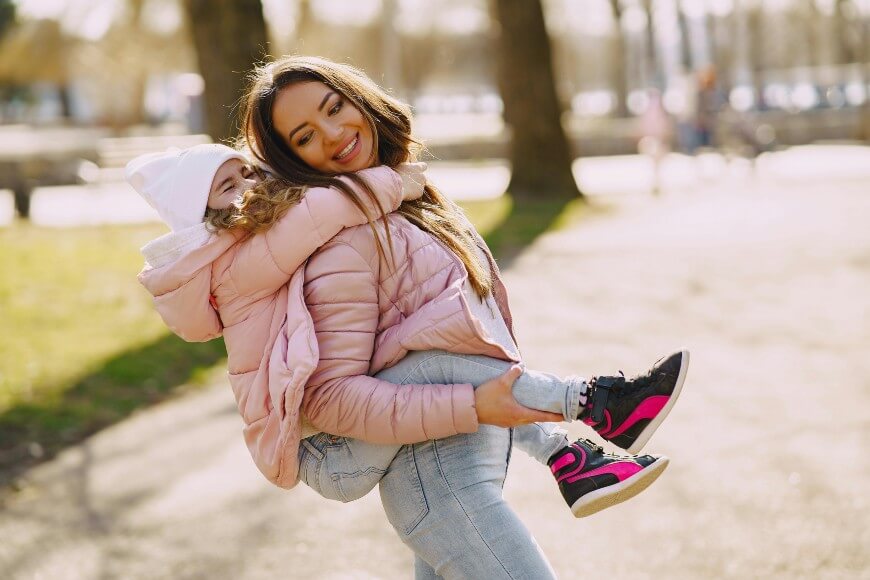 A woman in a pink jacket carries a smiling child on her back outdoors. Both are wearing light jackets and jeans. The child wears a white hat and sneakers. It’s a sunny day in a park with trees and blurred people in the background.