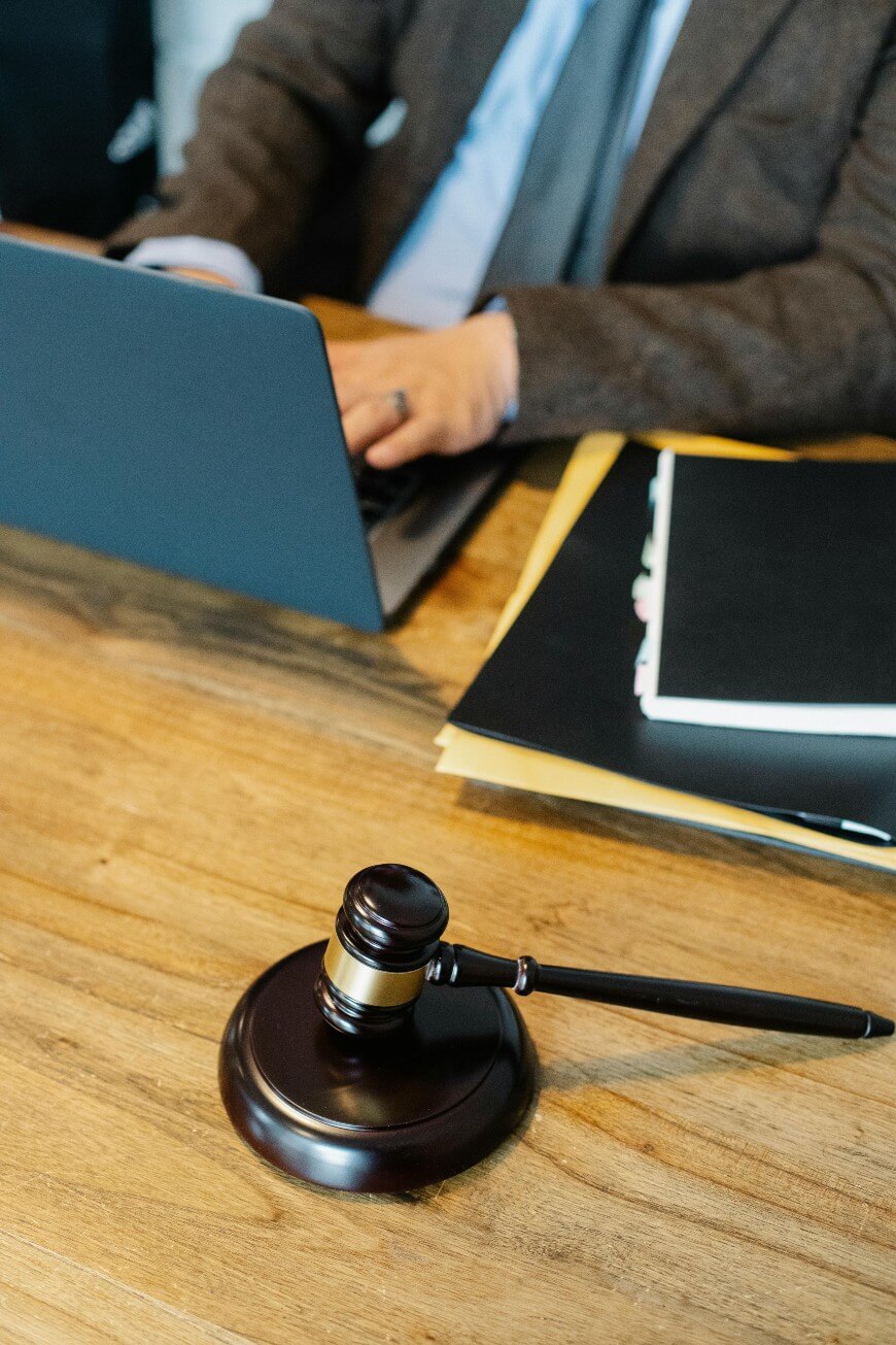A person in a suit types on a laptop at a wooden desk with a gavel and legal folders, suggesting a legal or judicial setting.
