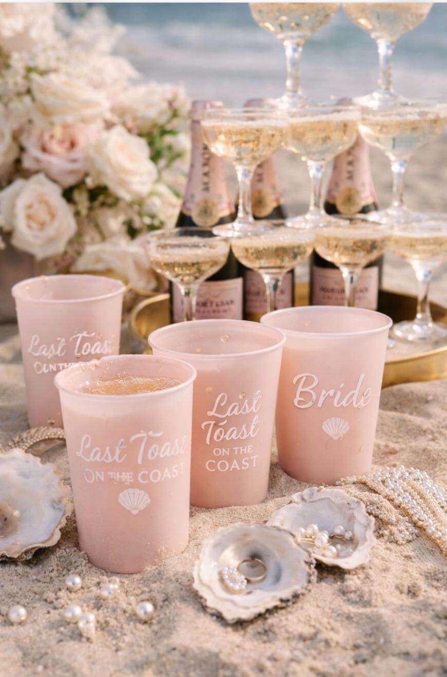 Pink cups with “Last Toast on the Coast” and “Bride” printed on them sit on sandy beach near oyster shells with pearls, champagne flutes, mini bottles, and a bouquet of white roses in the background.