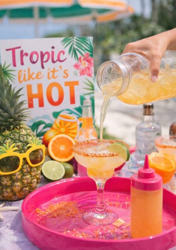A tropical-themed setup with a sign saying “Tropic like it’s HOT,” a pineapple with yellow sunglasses, and a hand pouring a yellow drink into a cocktail glass on a pink tray, surrounded by citrus fruits and drink bottles.