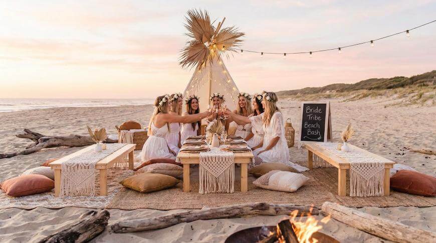 Five women in white dresses and flower crowns toast at a boho beach picnic with a teepee, low wooden tables, cushions, and a sign reading Bride Tribe Beach Bash at sunset. A fire pit and string lights add warmth.