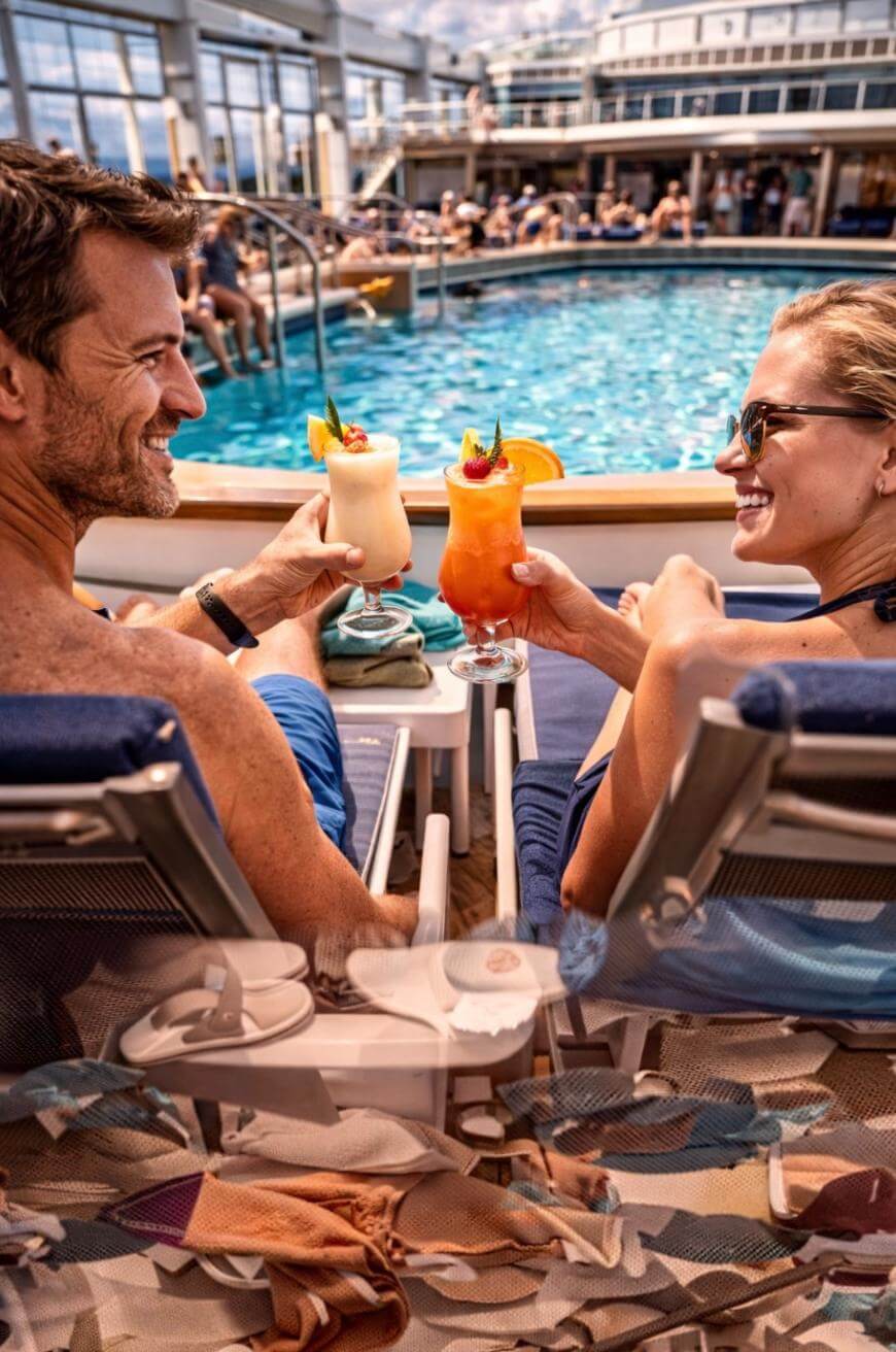 A man and woman relax on lounge chairs by a pool, smiling and toasting with colorful tropical drinks. People swim and relax in the background under a sunny, glass-covered area, suggesting a cruise ship or resort.