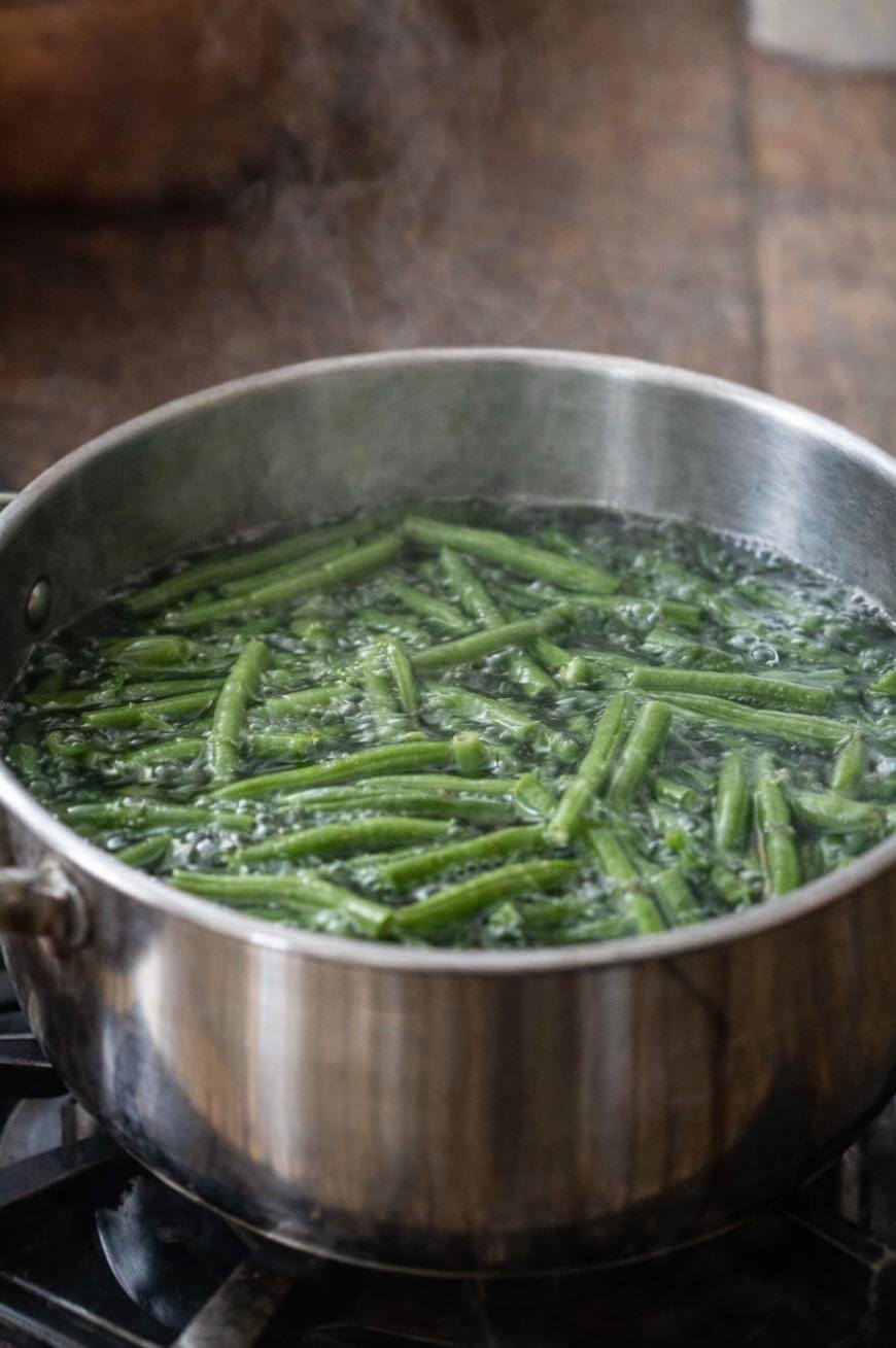 Green beans boiling in water inside a stainless steel pot on a stovetop, with steam rising from the pot.