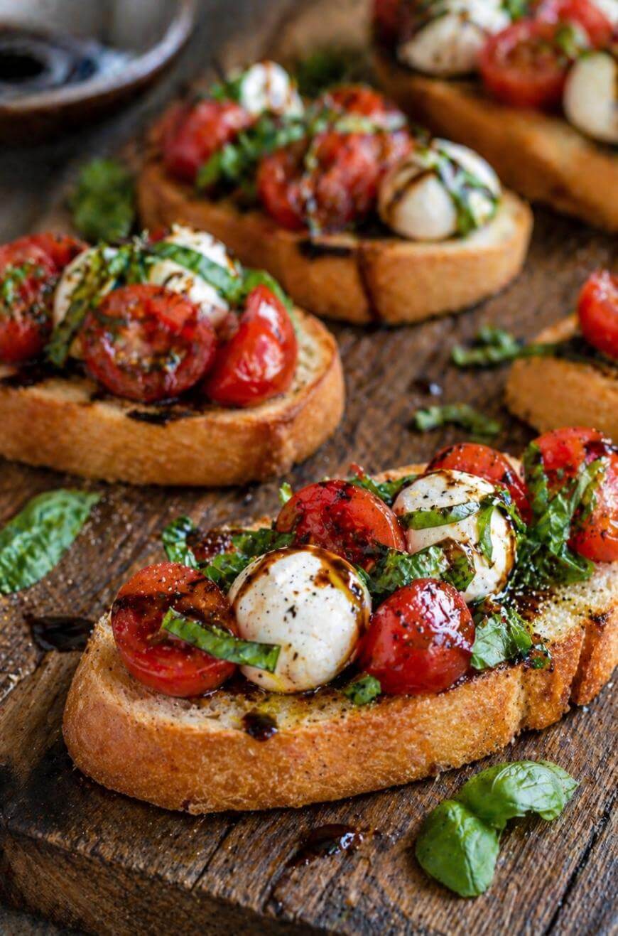 Slices of toasted bread topped with cherry tomatoes, mozzarella balls, fresh basil, and a drizzle of balsamic glaze, arranged on a rustic wooden board.