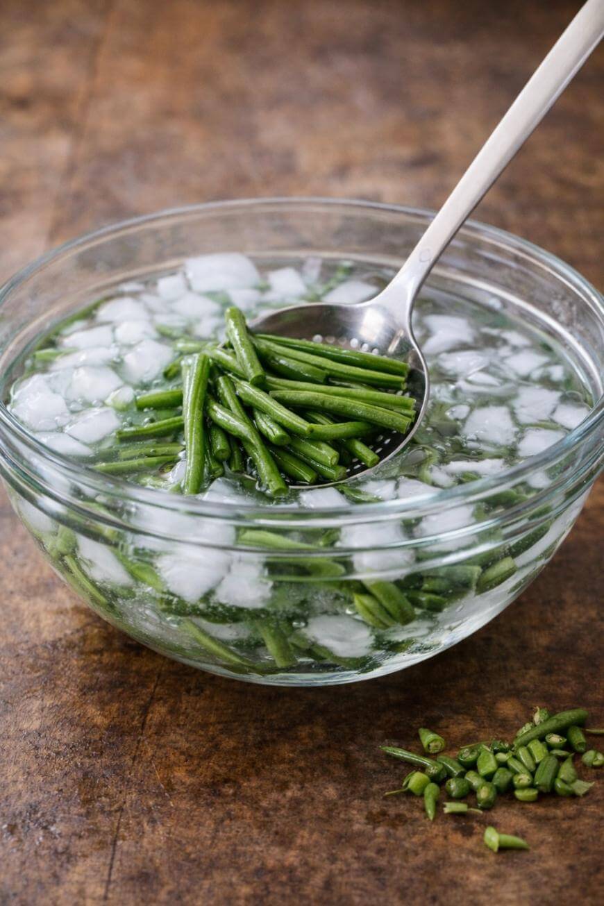 A glass bowl filled with ice water and green beans sits on a wooden surface. A metal spoon lifts some green beans out of the bowl. Chopped green bean pieces are scattered on the table nearby.