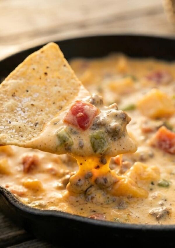 A tortilla chip dipped into a skillet of creamy queso dip with ground beef, tomatoes, and green peppers. The cheesy dip stretches from the skillet to the chip.