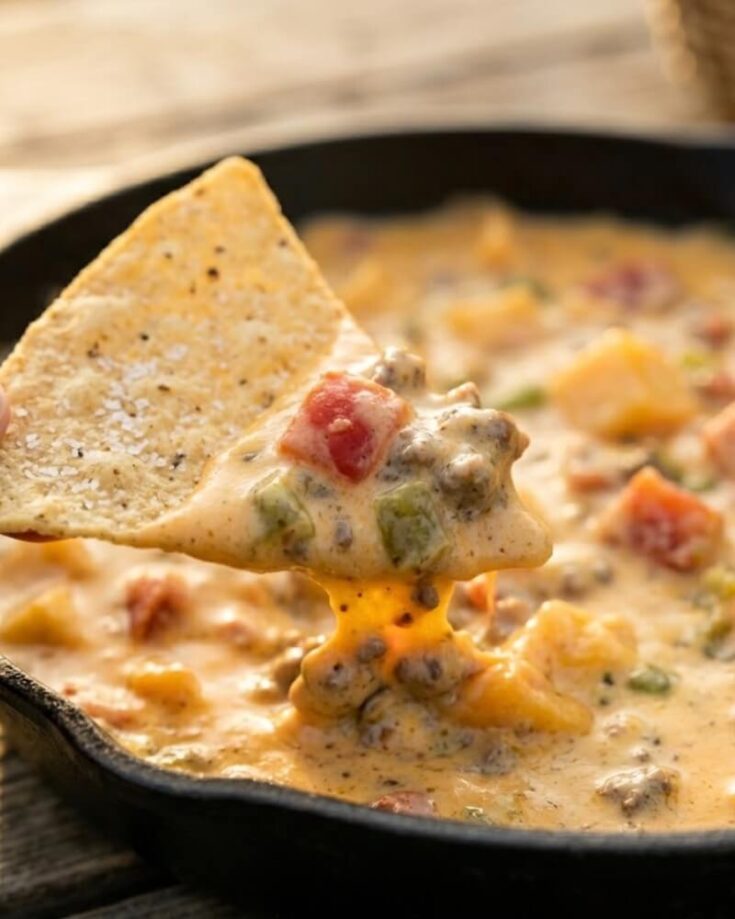 A tortilla chip dipped into a skillet of creamy queso dip with ground beef, tomatoes, and green peppers. The cheesy dip stretches from the skillet to the chip.