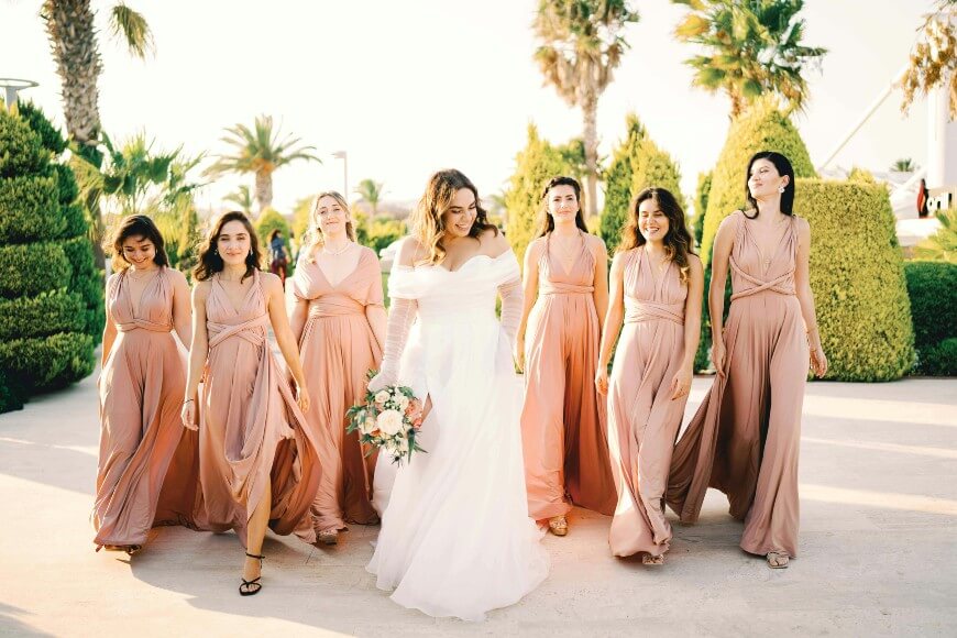 A bride in a white dress holding a bouquet walks outdoors with six bridesmaids in matching blush pink dresses, all smiling and surrounded by lush greenery and palm trees.