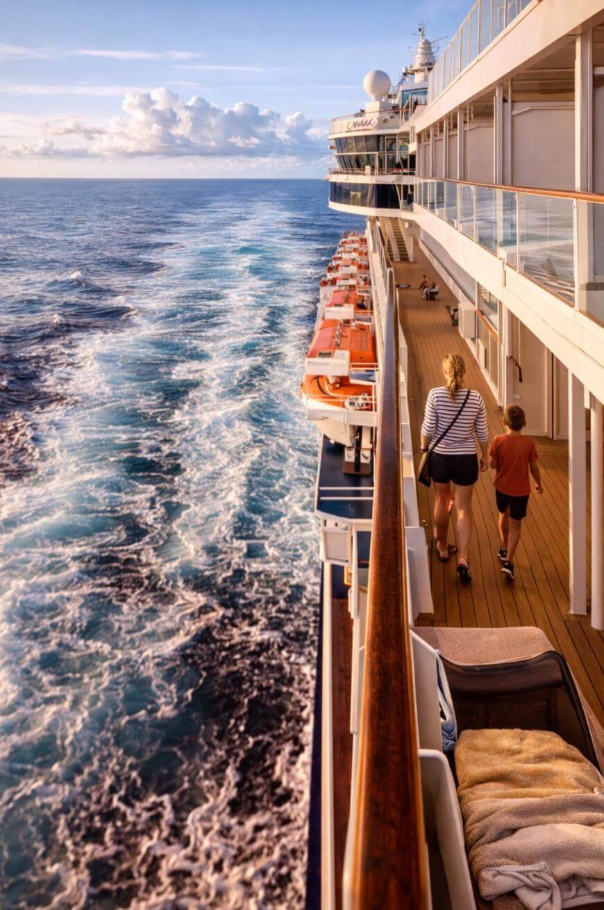A woman and a child walk along the deck of a cruise ship, with orange lifeboats beside them and the ship’s wake trailing in the blue ocean under a partly cloudy sky.
