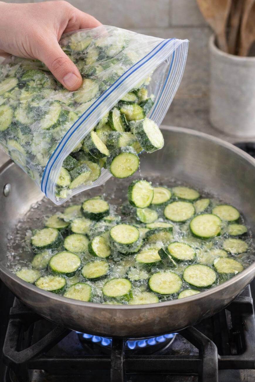 A hand pours frozen sliced zucchini from a resealable plastic bag into a stainless steel skillet on a lit stovetop. The zucchini is covered in ice crystals, and the pan is starting to sizzle.